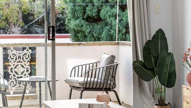 A room with a balcony, a chair, a table with a book, a potted plant, and a decorative wall piece. - Cosy & Convenient Place in Inner West Area (no parking) (Strathfield)