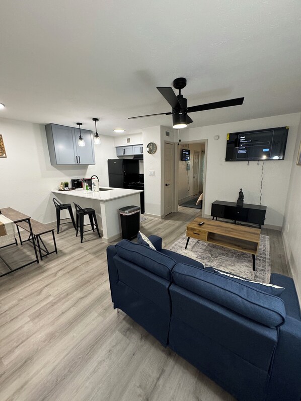 A modern living room with a blue sofa, a wooden coffee table, and a kitchen area with grey cabinets and a black refrigerator.