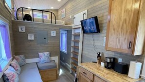 A compact kitchen with wooden cabinets, a built-in seating area, a television, and a small table with a tablet. - The Mermaid at Tiny House Siesta (Sarasota)