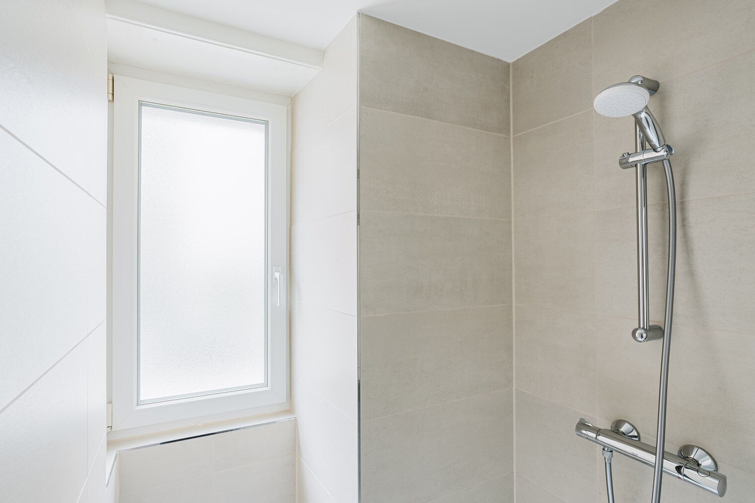 A shower with a handheld showerhead and a wall-mounted faucet, a window with frosted glass, and a corner with beige tiles.