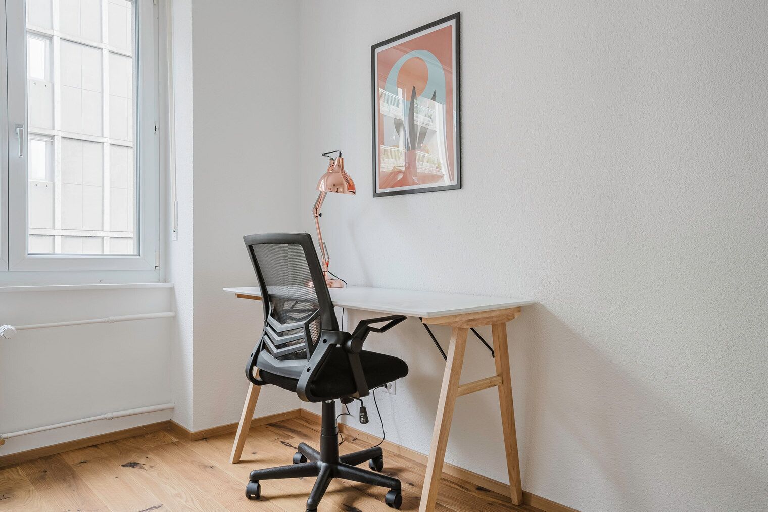 A small office space with a white desk, a black office chair, a framed picture on the wall, and a window with blinds.