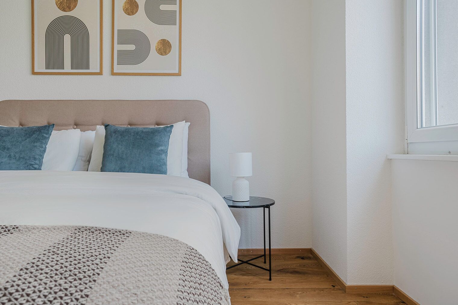 A neatly arranged bedroom with a bed, pillows, a bedside table with a lamp, and two framed artworks on the wall.