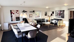 Una sala de estar moderna con comedor, que incluye un sofá, una mesa de centro y una mesa de comedor con sillas. Hay estantes en la pared con objetos decorativos y un ventanal que proporciona luz natural.