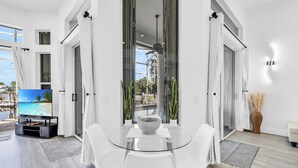 A modern living room with a glass dining table, white chairs, a flat-screen TV, and a view of a pool and palm trees. - Spacious waterfront Oasis with heated pool. (Marco Island)