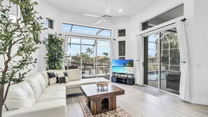 A spacious living room with a white leather sofa, a wooden coffee table, and a large flat-screen TV. The room has large windows offering a view of a pool and palm trees. - Spacious waterfront Oasis with heated pool. (Marco Island)
