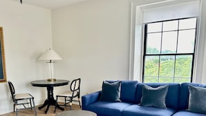 A living room with a blue sectional sofa, two round tables, a patterned rug, and a window with a view of greenery. - Modern Suite on the River (Port Hope)