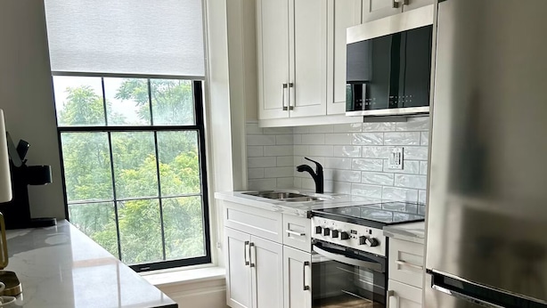 A modern kitchen with white cabinets, a stainless steel refrigerator, a built-in oven, and a window with a view of greenery. - Modern Suite on the River (Port Hope)