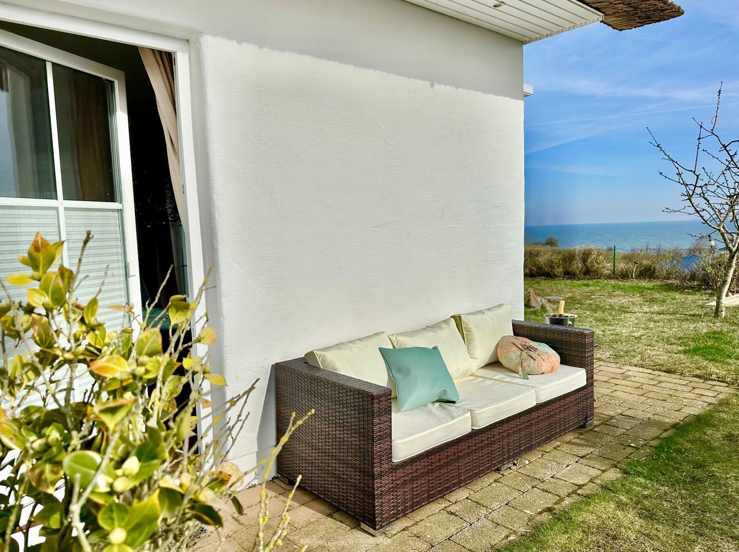 A wicker sofa with cushions and a white wall behind it, set on a paved area with a view of the sea.