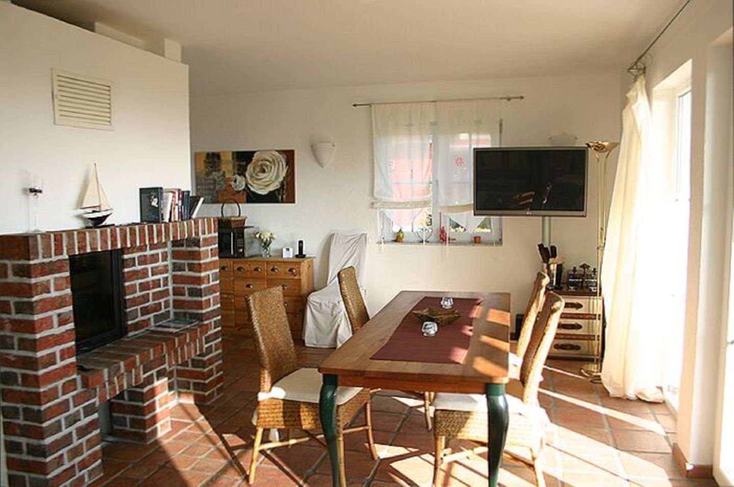 Ein Essbereich mit Kamin aus Ziegelsteinen, Korbstühlen, einem Holztisch und einem an der Wand montierten Fernseher.