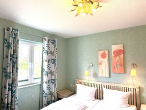 A bedroom with a bed, curtains, a nightstand, a lamp, and two wall art pieces. - Close to the sea, quiet location, use of swimming pool and sauna - Reethaus Göhren FeWo 01 (Göhren)