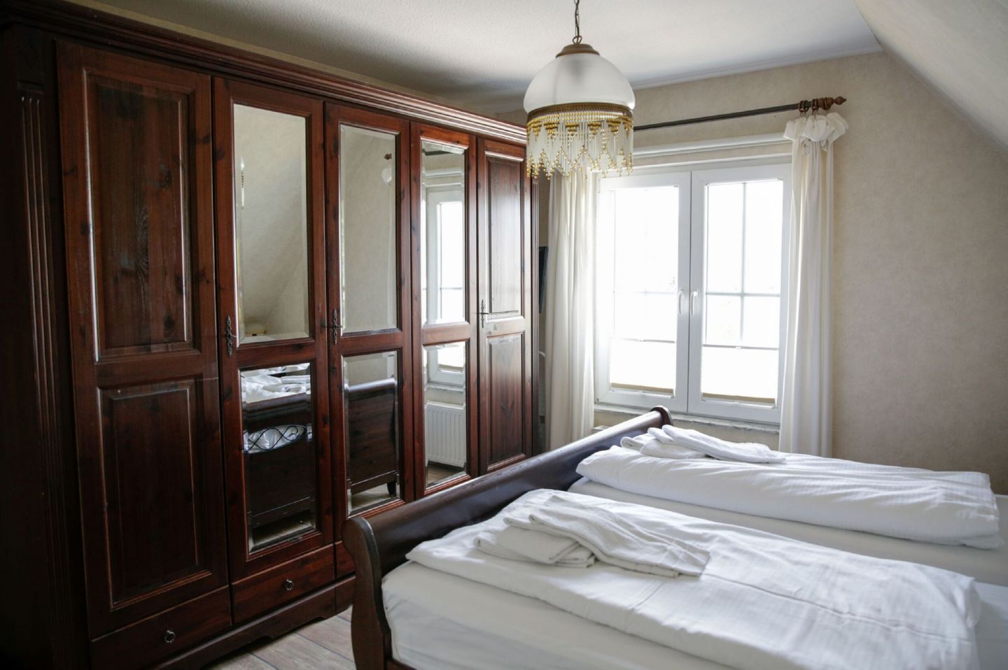 A bedroom with a large wooden wardrobe, a bed with white linens, and a window with curtains.