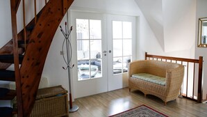 Une pièce avec un escalier en bois, un canapé en osier, un lampadaire et une vue sur l’extérieur, visible à travers une porte.