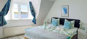 Un dormitorio con cama, ventana con cortinas, radiador, un cuadro en la pared y una mesita de noche con lámpara.
