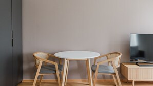 Ein Zimmer mit einem runden weißen Tisch, zwei Korbstühlen mit grauen Kissen, einem dunklen Kleiderschrank und einem Fernseher auf einer Holzkommode.