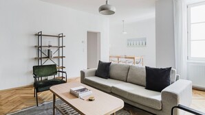 A modern living room with a sofa, a coffee table, a bookshelf, and a dining area in the background. - Upscale Neubau 3BR w/ Elevator & W/D, by Blueground (Vienna)