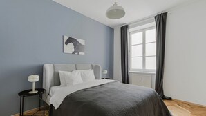 A modern bedroom with a grey headboard, a large window with curtains, a bedside table with a lamp, and a wall-mounted artwork. - Upscale Neubau 3BR w/ Elevator & W/D, by Blueground (Vienna)