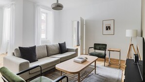A modern living room with a sofa, coffee table, and chairs. There is a lamp, a framed picture on the wall, and a window with curtains. - Upscale Neubau 3BR w/ Elevator & W/D, by Blueground (Vienna)