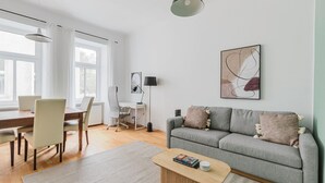 Een moderne woonkamer met een grijze bank, een eettafel met stoelen, een staande lamp en een schilderij aan de muur.