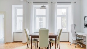 Een eetruimte met een houten tafel en zes stoelen, grote ramen met witte gordijnen en een plafondlamp.