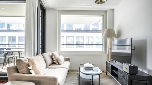 A modern living room with a sofa, coffee table, TV, and a large window offering a view of the cityscape.