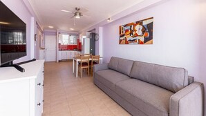 Un salon avec un canapé gris, une table à manger avec des chaises, un coin cuisine avec un réfrigérateur et un ventilateur de plafond.
