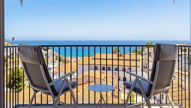 Un balcon avec deux chaises et une petite table, d’où l’on peut admirer une ville côtière et la mer.