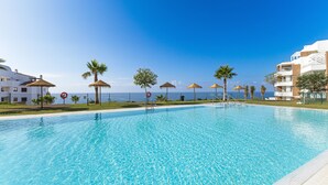 A clear blue swimming pool with a tiled deck, surrounded by palm trees and umbrellas, with a view of the sea and buildings in the background. - Isea Calaceite, 2 bedroom (Llanos, Los (Torrox-Costa))