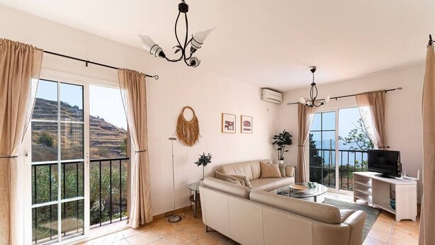 A spacious living room with a sofa, a coffee table, and a TV. There are large windows with curtains, a chandelier, and a view of the outdoors. - Private villa in Tamango Hill (Torrox)