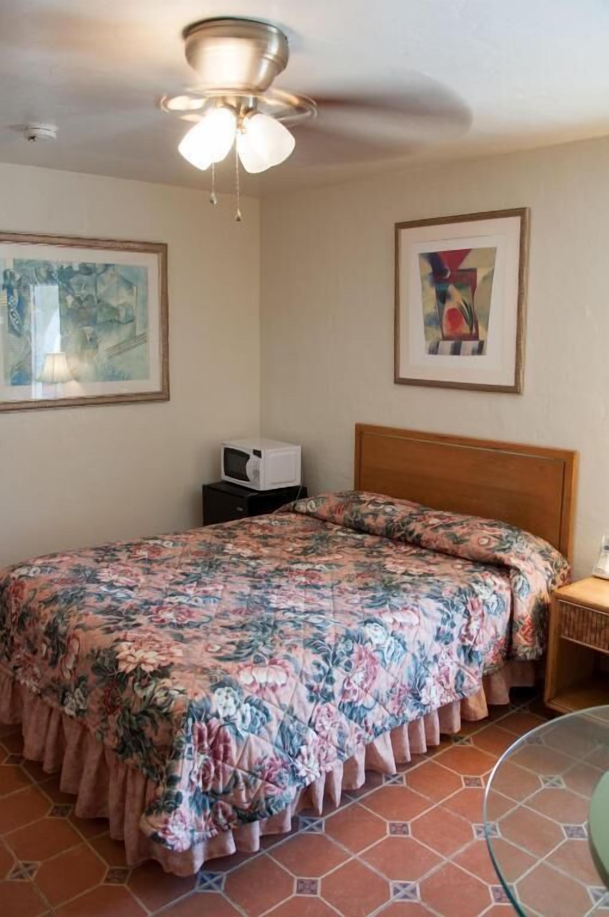 A bedroom with a floral bedspread, a wooden headboard, a ceiling fan, a microwave on a small table, and two framed artworks on the walls.