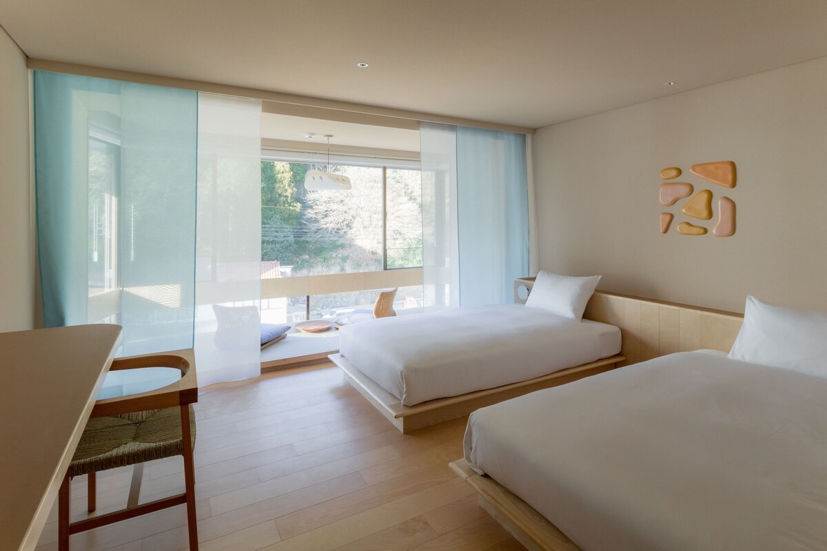 A modern hotel room with two beds, a desk, and a large window offering a view of greenery.