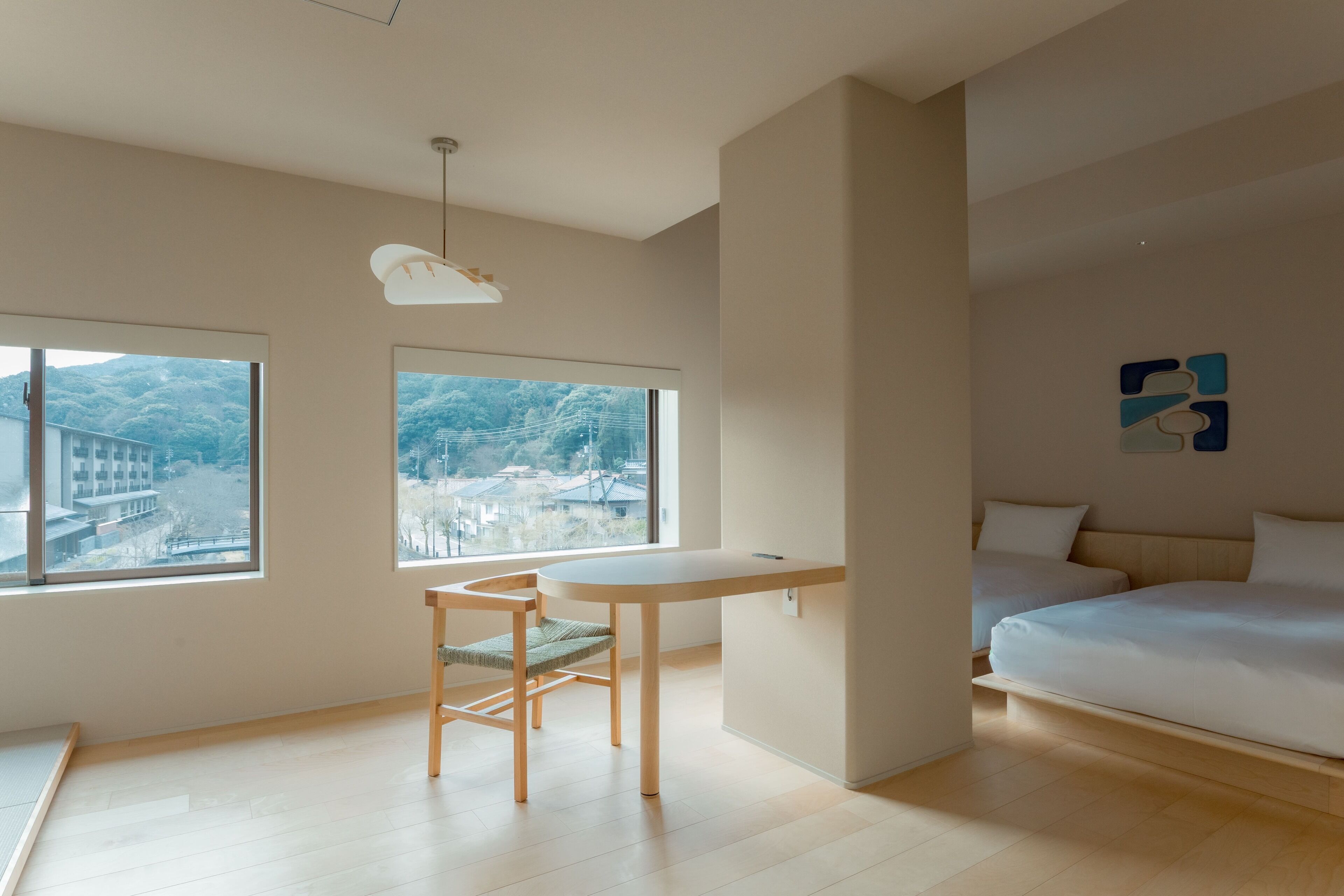 Quarto moderno com uma cama, escrivaninha e duas janelas com vista para uma ponte e colinas.