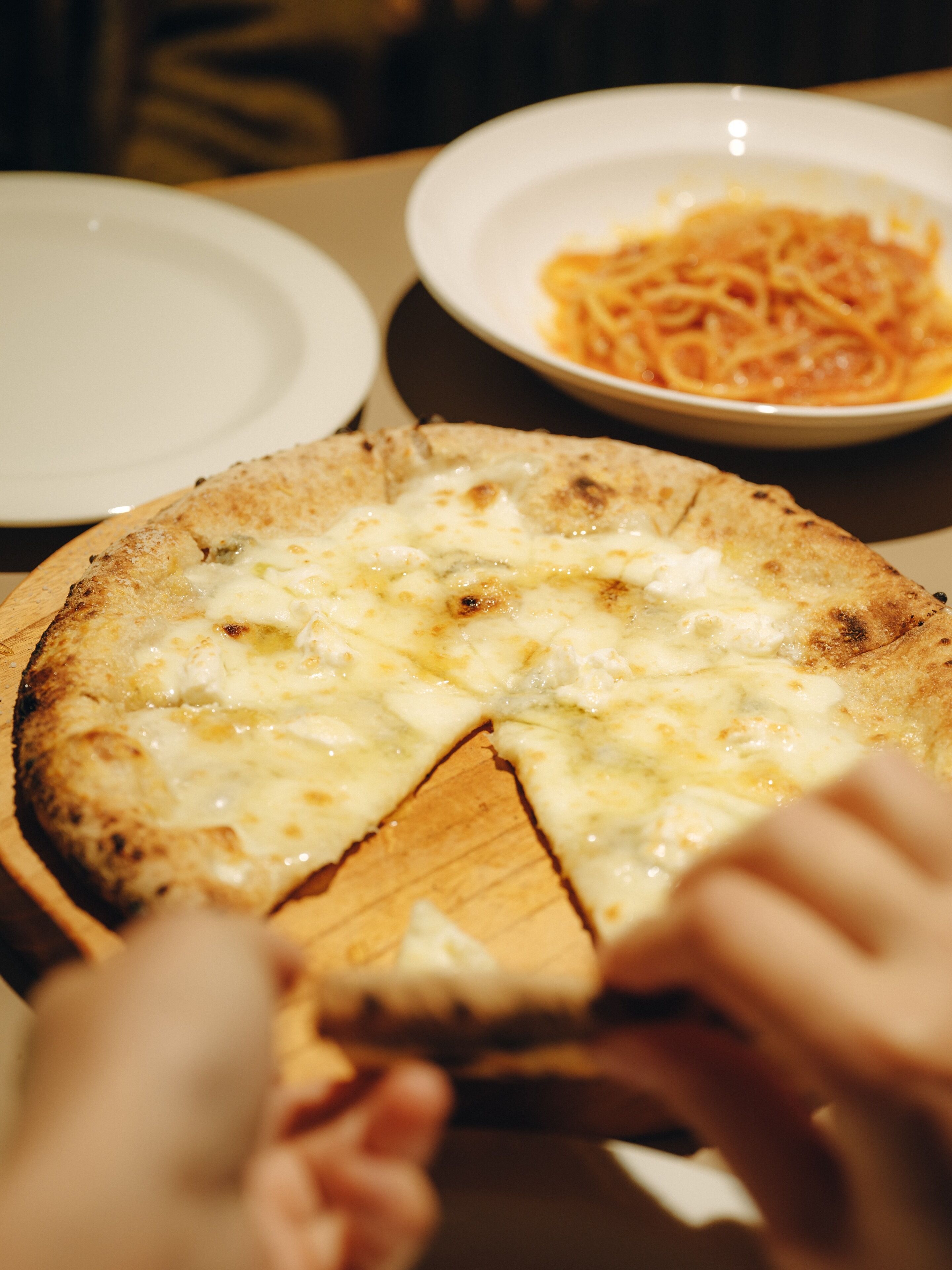 Une pizza sur une planche en bois, avec une part manquante, et une assiette de spaghettis en arrière-plan.