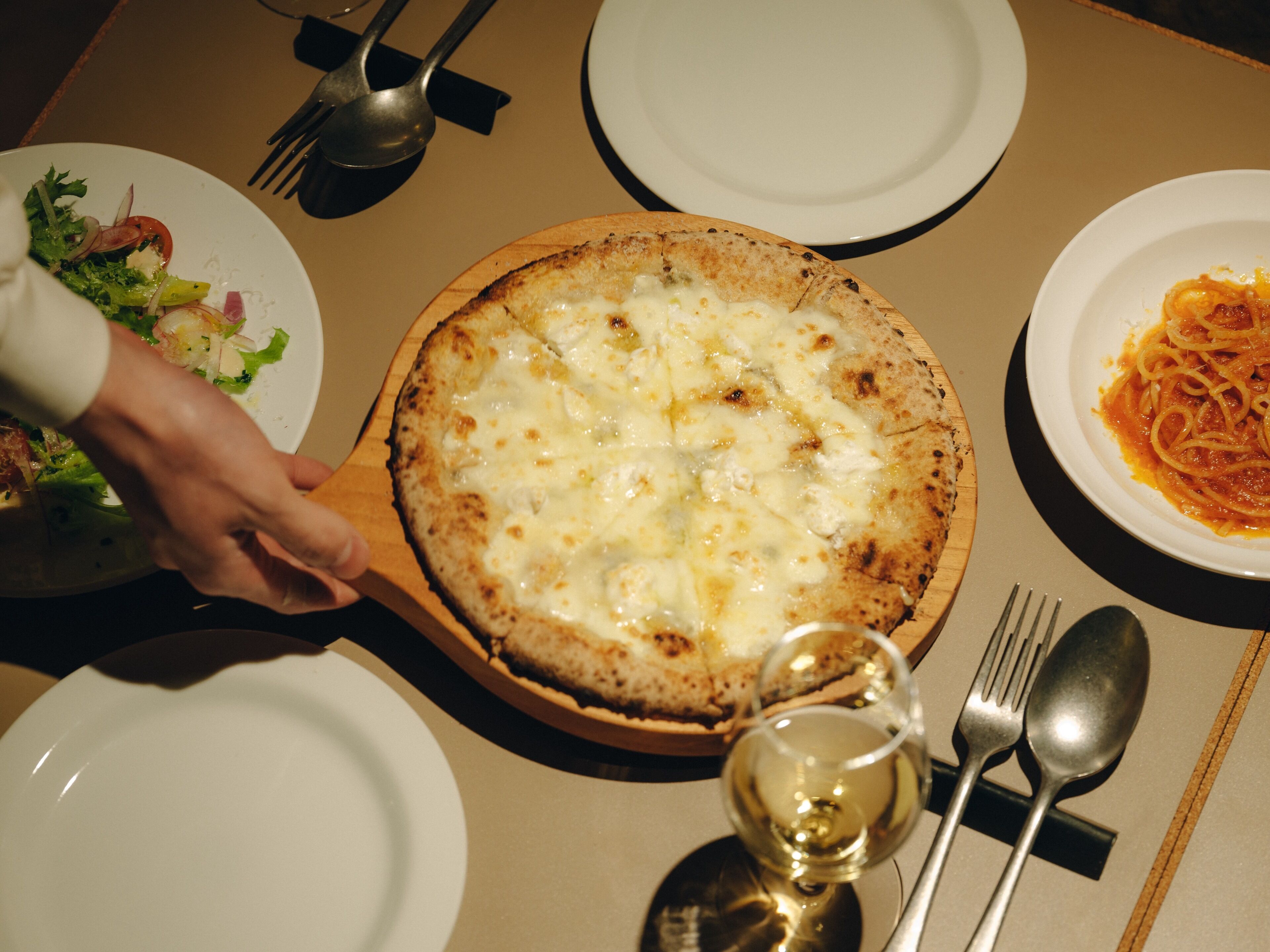 A dining table with a pizza, a plate of pasta, and a glass of wine.