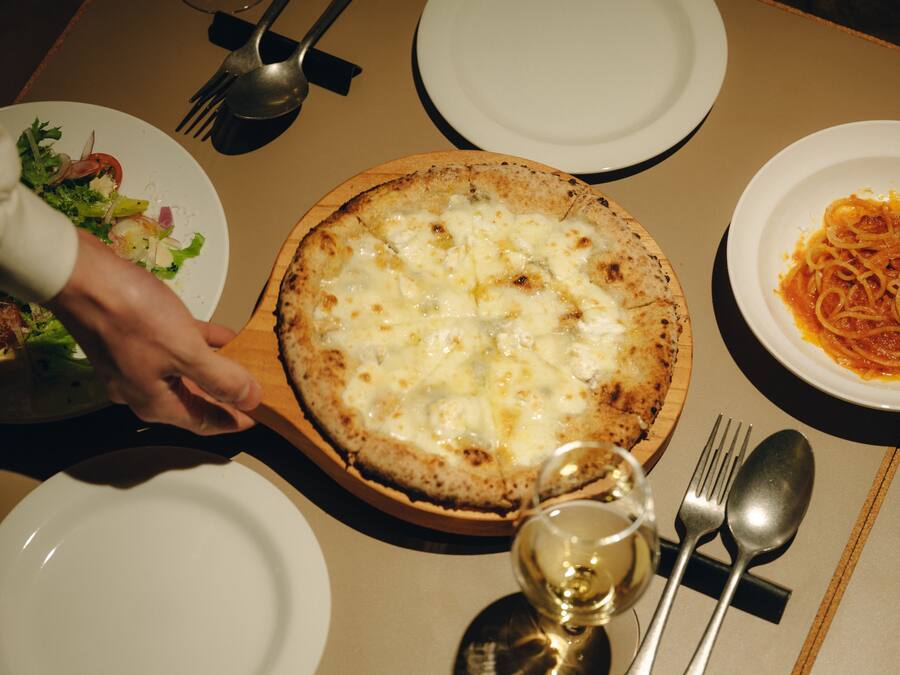 A dining table with a pizza, a plate of pasta, and a glass of wine.