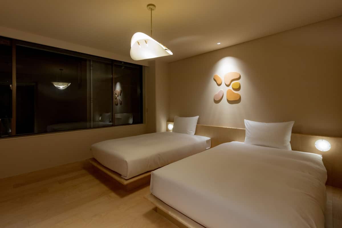 A modern bedroom with two beds, a large window, and a unique wall art piece.