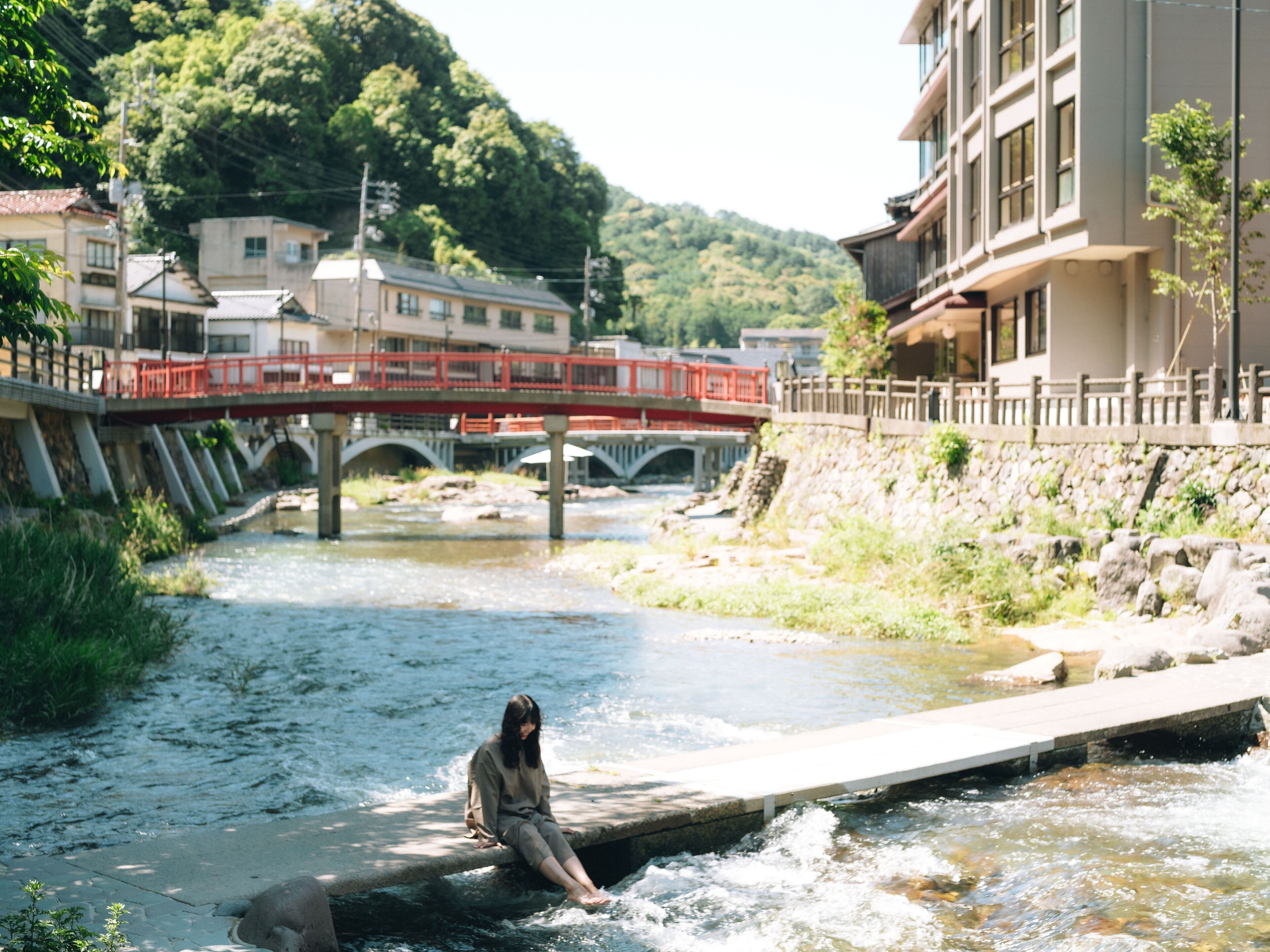 A woman sitting on a concrete ledge by a river, with a bridge and buildings in the background.