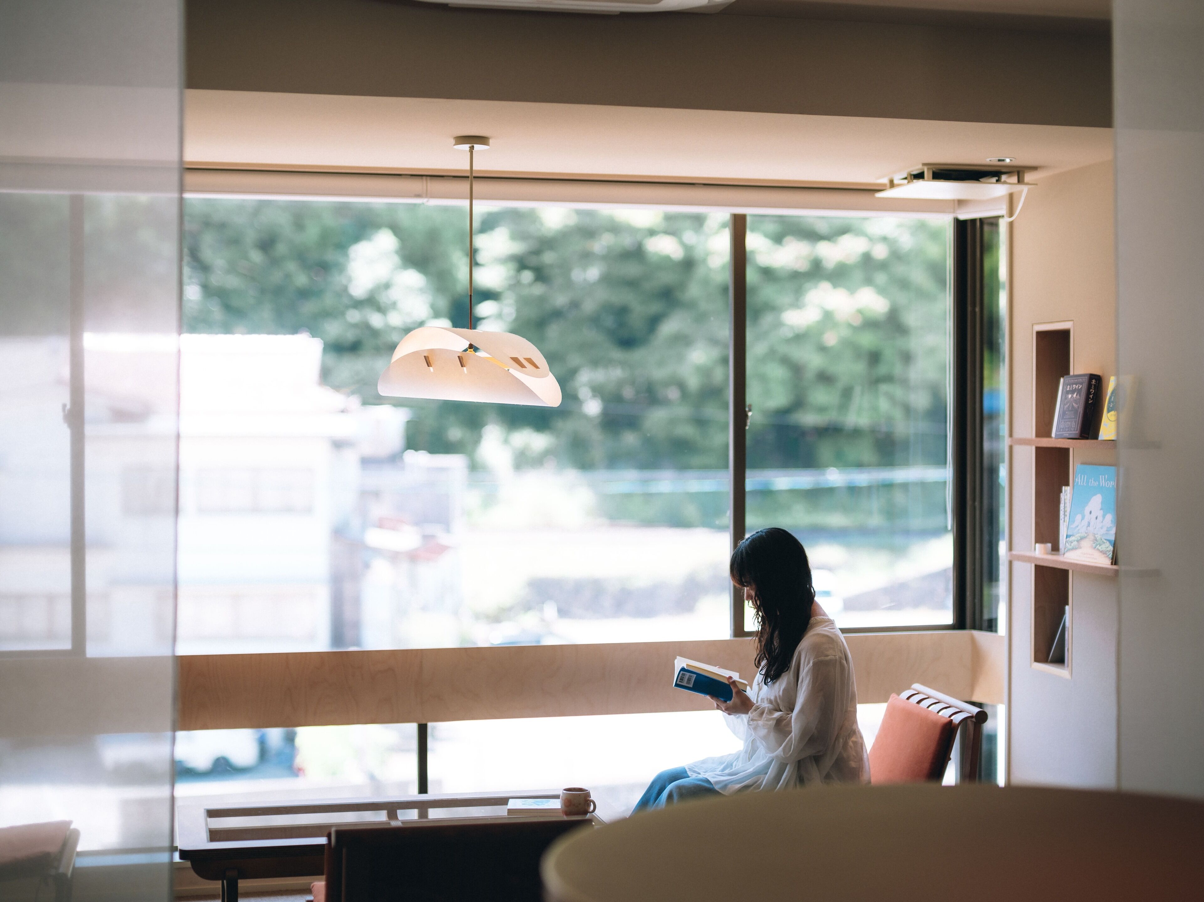 Una persona sentada junto a una ventana leyendo un libro en una habitación con un ventanal grande, una lámpara colgante y una estantería.