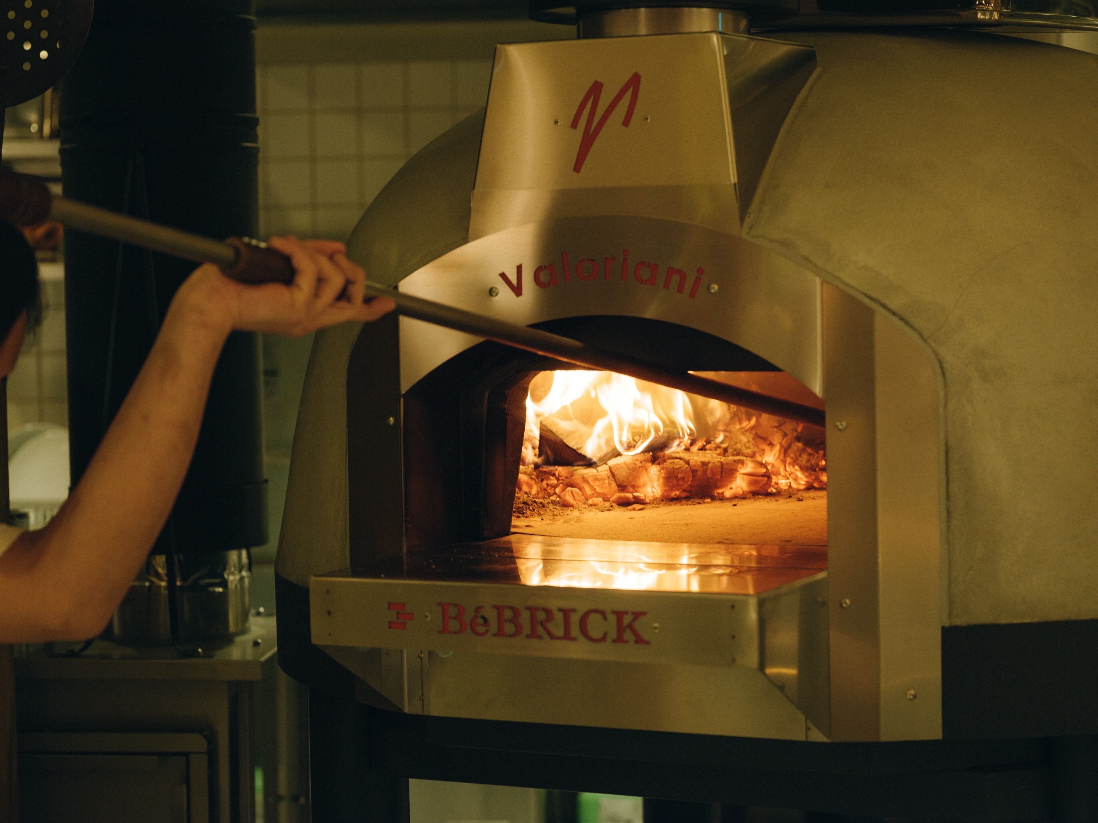 A person using a long-handled tool to open a metal oven door with a fire inside.