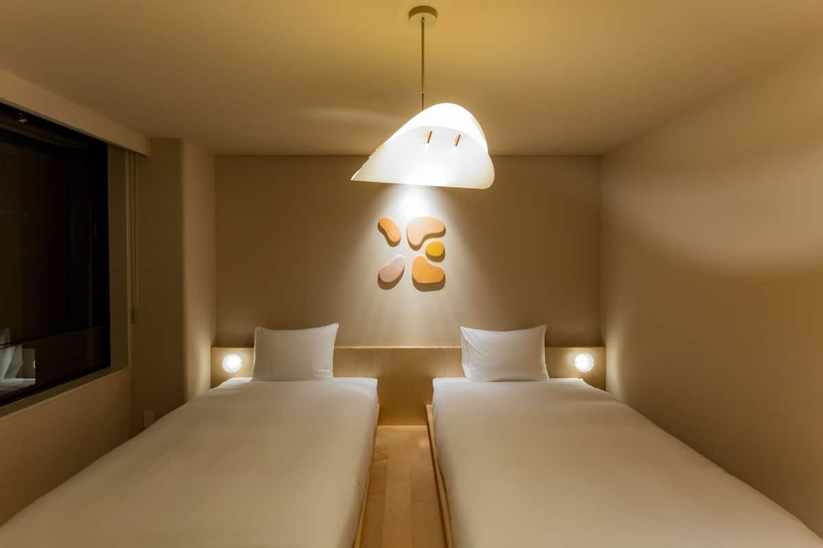 A hotel room with two beds, a wall-mounted light fixture, and a window on the left.