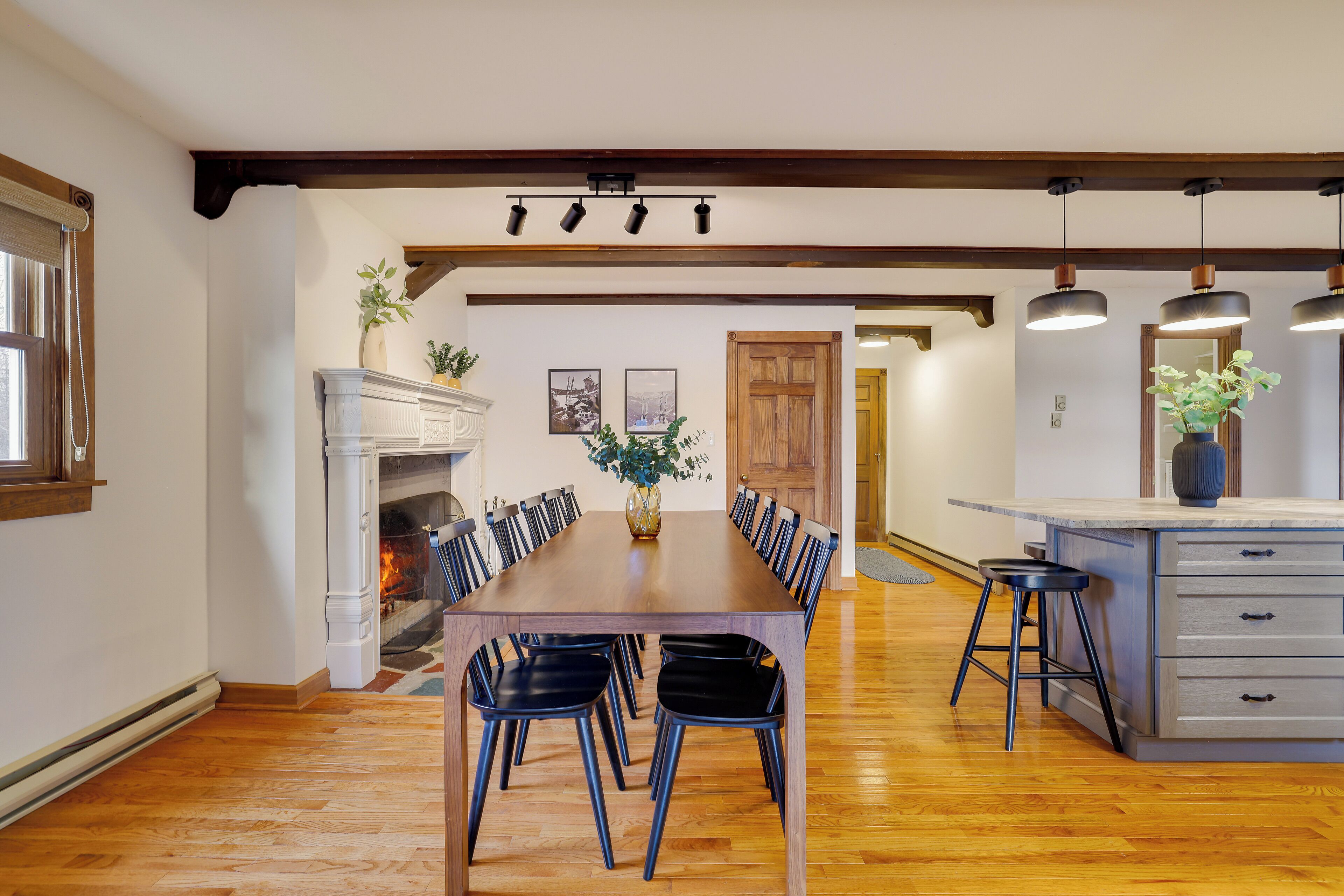 Un comedor con una larga mesa de madera, sillas negras, chimenea y una zona de cocina con un taburete de bar.