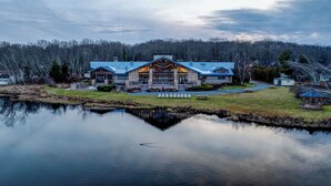 A large, two-story lodge with a prominent entrance and a reflection in the water in front. - Arrowhead Premier Lakefront w/Sauna, Hot Tub, Firepit, Game Room, Amenities, (Pocono Lake)