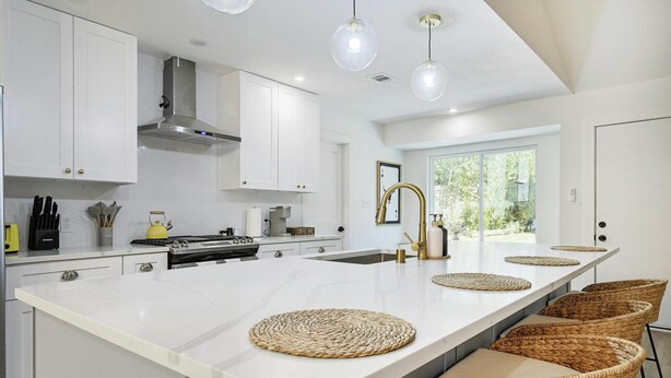 A modern kitchen with white cabinetry, a central island with woven placemats, and a large window providing natural light. - Designer Home,  Sleep 8,  Ask us about FREE add-ons (Austin)