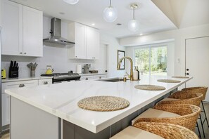 A modern kitchen with white cabinetry, a central island with woven placemats, and a large window providing natural light. - Designer Home,  Sleep 8,  Ask us about FREE add-ons (Austin)