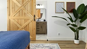 A bedroom with a wooden sliding door, a bed with a blue bedspread, a large potted plant, and a kitchen area with cabinets and a countertop. - Designer Home,  Sleep 8,  Ask us about FREE add-ons (Austin)