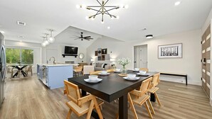 A modern kitchen with a central island, wooden dining table, and chairs. There is a ceiling fan, recessed lighting, and a large window providing natural light. - Designer Home,  Sleep 8,  Ask us about FREE add-ons (Austin)