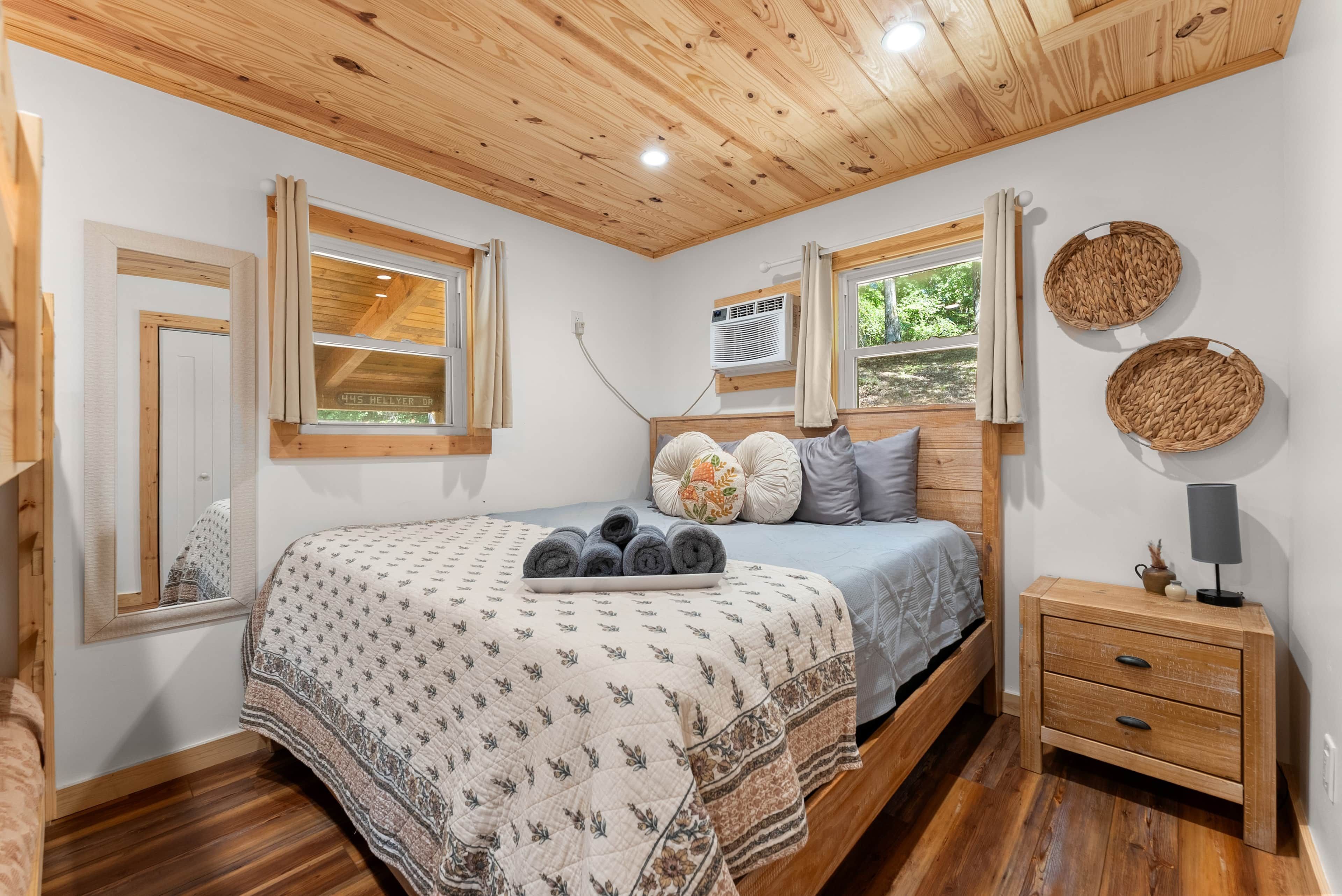 A bedroom with a wooden ceiling, a bed with a floral quilt, a bedside table with a lamp, and a window with a view of greenery.