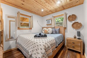 A bedroom with a wooden ceiling, a bed with a floral quilt, a bedside table with a lamp, and a window with a view of greenery. - River Retreat Cabin (Cleveland)