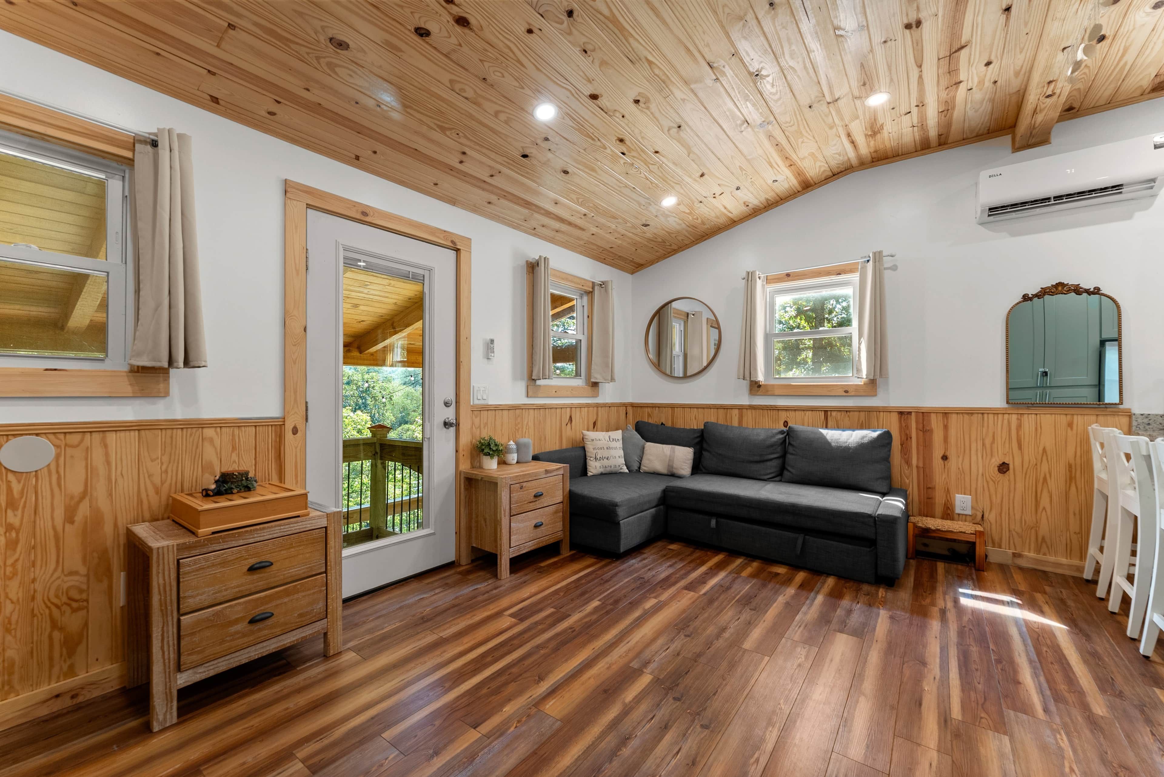 A room with wooden flooring, a sofa, a wooden cabinet, and a door leading to an outdoor area.