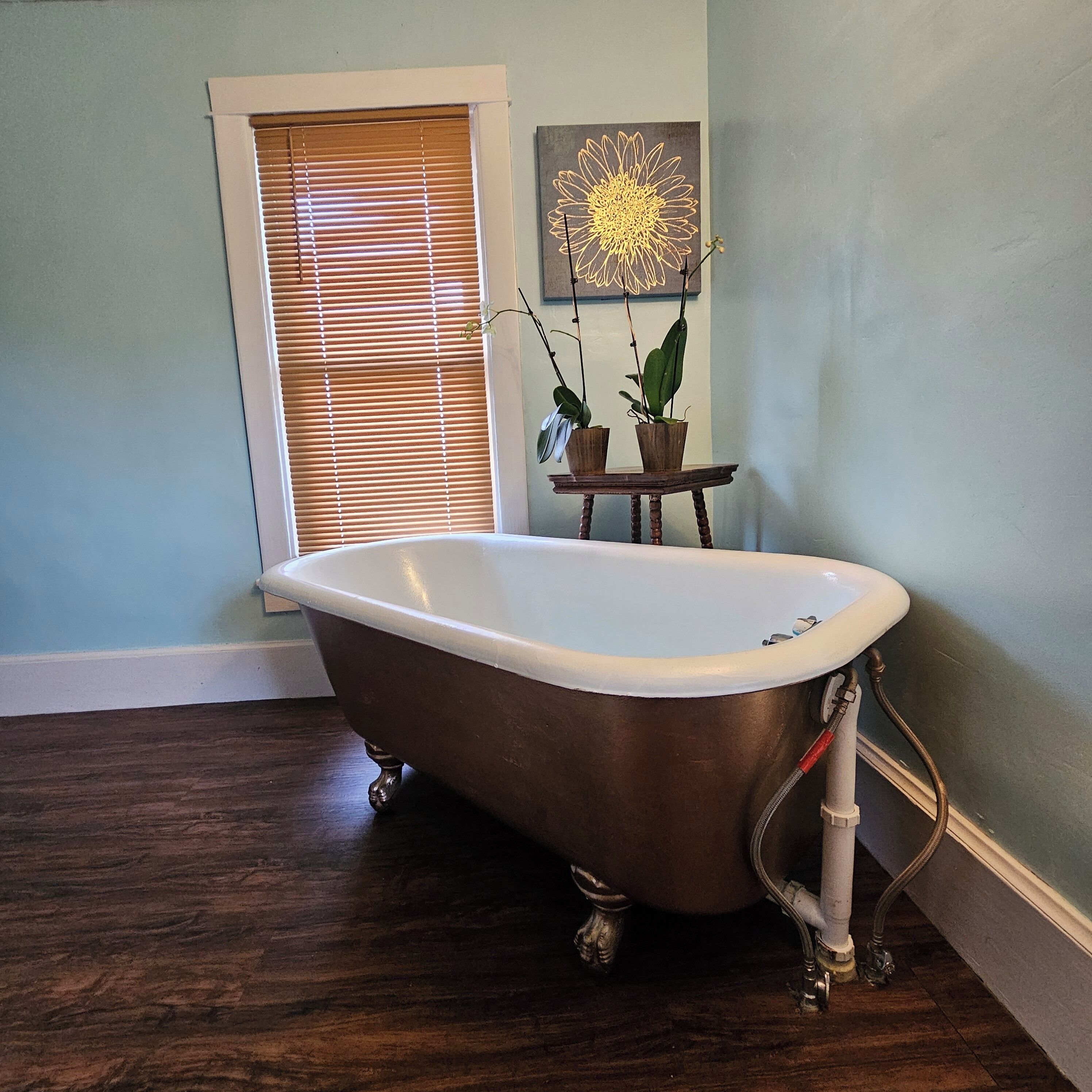 A bathroom with a freestanding bathtub, wooden flooring, a window with blinds, and a small table with two potted plants.