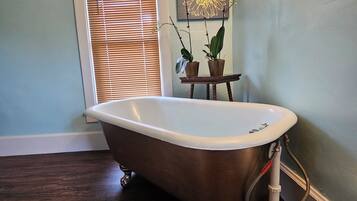 A bathroom with a freestanding bathtub, wooden flooring, a window with blinds, and a small table with two potted plants.